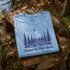 Folded blue t-shirt with nature-themed design on a wooden surface with pinecones and leaves.