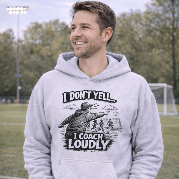 man wearing a grey hoodie with “I Don’t Yell I Coach Loudly” soccer coach design featuring coach pointing on sideline with players in background
