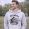 man wearing a grey hoodie with “I Don’t Yell I Coach Loudly” soccer coach design featuring coach pointing on sideline with players in background