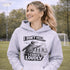 Woman wearing a grey hoodie with “I Don’t Yell I Coach Loudly” soccer coach design featuring coach pointing on sideline with players in background