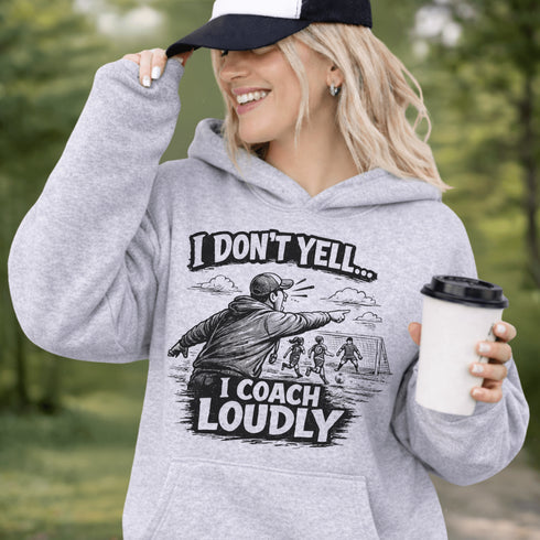Woman wearing a grey hoodie with “I Don’t Yell I Coach Loudly” soccer coach design featuring coach pointing on sideline with players in background