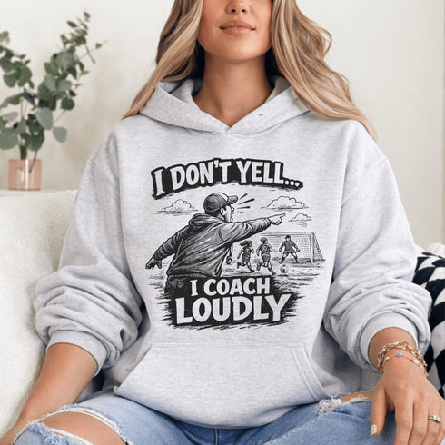 Woman wearing a grey hoodie with “I Don’t Yell I Coach Loudly” soccer coach design featuring coach pointing on sideline with players in background