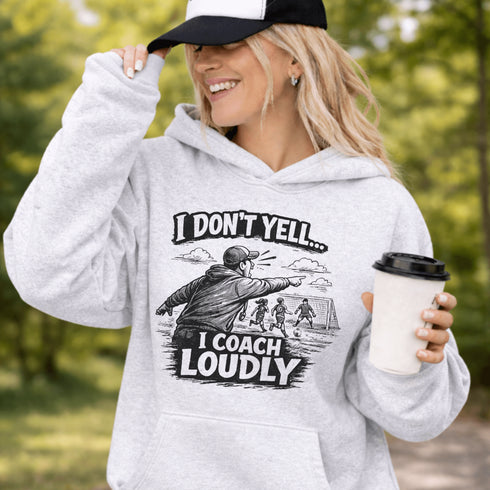 Woman wearing a grey hoodie with “I Don’t Yell I Coach Loudly” soccer coach design featuring coach pointing on sideline with players in background