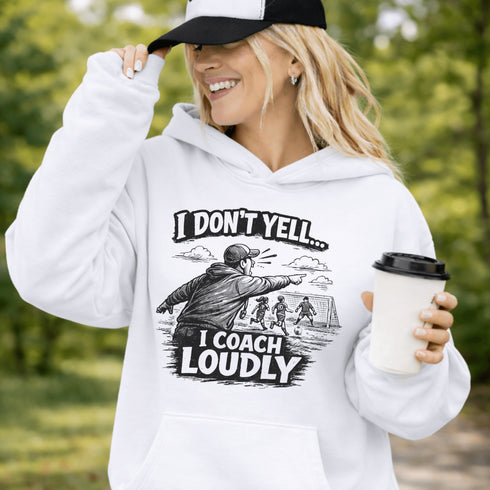 Woman wearing a grey hoodie with “I Don’t Yell I Coach Loudly” soccer coach design featuring coach pointing on sideline with players in background