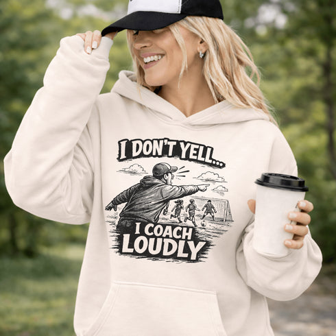Woman wearing a grey hoodie with “I Don’t Yell I Coach Loudly” soccer coach design featuring coach pointing on sideline with players in background