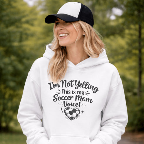 Woman wearing a hoodie that says “I’m Not Yelling This Is My Soccer Mom Voice” with a soccer ball heart design, standing on a soccer field