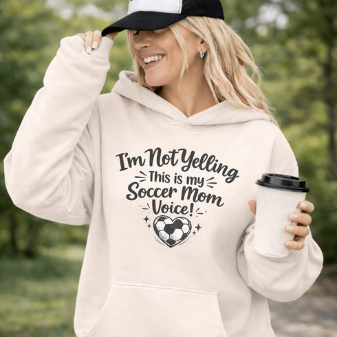 Woman wearing a hoodie that says “I’m Not Yelling This Is My Soccer Mom Voice” with a soccer ball heart design, standing on a soccer field