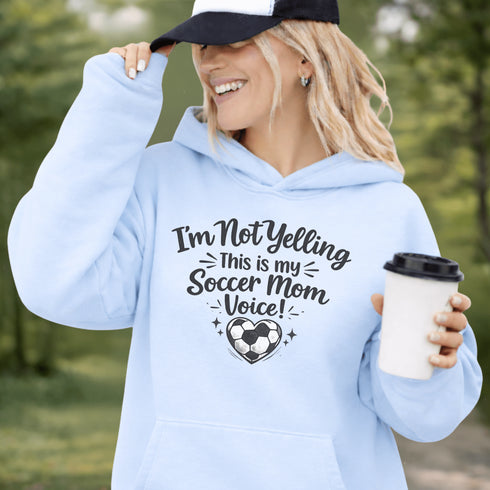 Woman wearing a hoodie that says “I’m Not Yelling This Is My Soccer Mom Voice” with a soccer ball heart design, standing on a soccer field