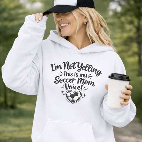 Woman wearing a hoodie that says “I’m Not Yelling This Is My Soccer Mom Voice” with a soccer ball heart design, standing on a soccer field