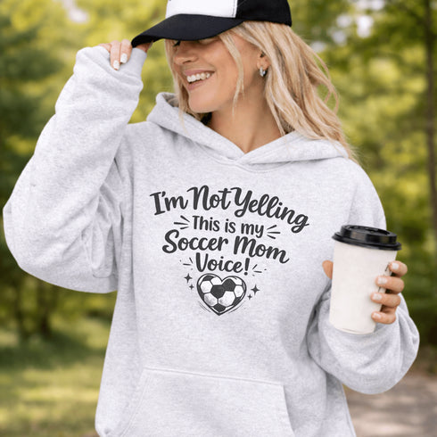 Woman wearing a hoodie that says “I’m Not Yelling This Is My Soccer Mom Voice” with a soccer ball heart design, standing on a soccer field
