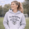 Woman wearing a grey hoodie that says “I’m Not Yelling This Is My Soccer Mom Voice” with a soccer ball heart design, standing on a soccer field