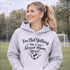 Woman wearing a grey hoodie that says “I’m Not Yelling This Is My Soccer Mom Voice” with a soccer ball heart design, standing on a soccer field
