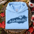 A light blue folded hoodie displayed on a rustic tree stump with autumn leaves and a plaid blanket around it. The hoodie features a dark blue graphic of Lake Ontario's map outline with the text 'it's Lake Ontario Eh!'.