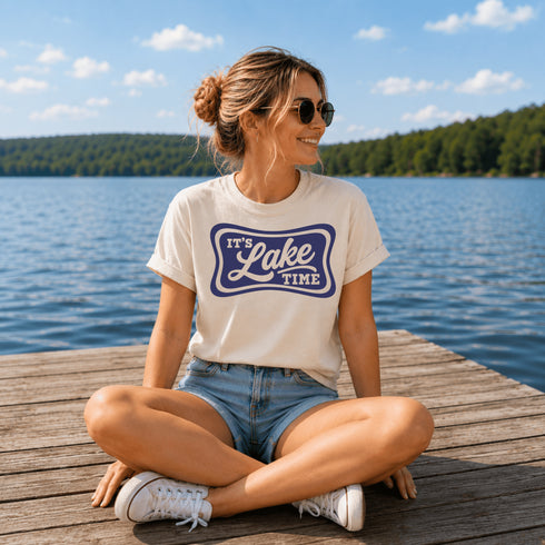 Woman sitting on a dock by a lake wearing a 'It's Lake Time' t-shirt.