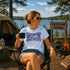 Woman sitting by a campfire at Falcon Lake, Manitoba, holding a mug with 'It's Lake Time' t-shirt.