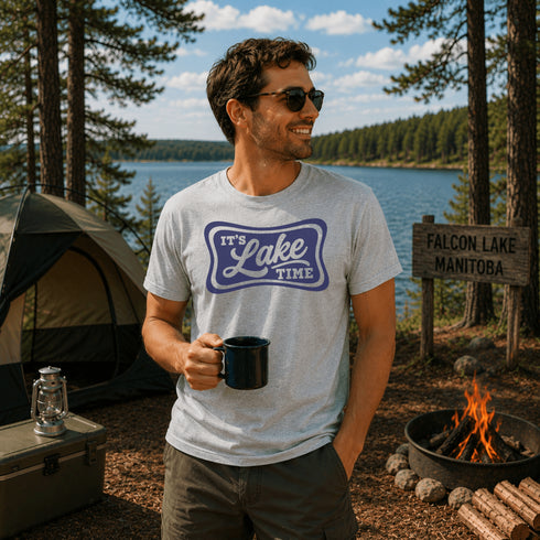 Man in a 'It's Lake Time' t-shirt by a campfire with a lake and forest in the background.