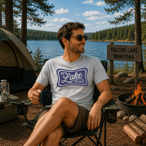 Man sitting by a campfire with a lake and forest in the background, wearing a 'It's Lake Time' t-shirt.