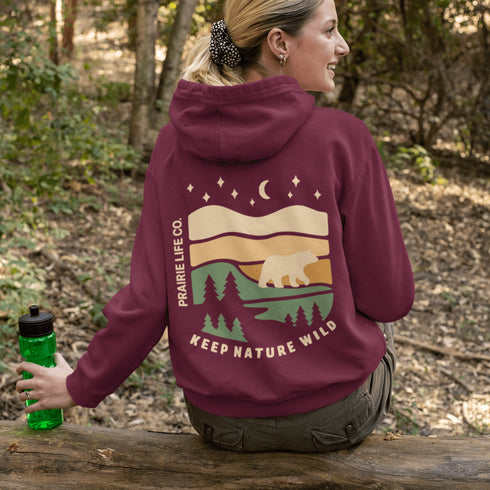 This graphic features a bear silhouette wandering through layered prairie hills, pine forests, and a serene night sky filled with stars and a crescent moon.