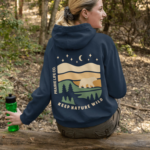 This graphic features a bear silhouette wandering through layered prairie hills, pine forests, and a serene night sky filled with stars and a crescent moon.