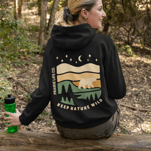 This graphic features a bear silhouette wandering through layered prairie hills, pine forests, and a serene night sky filled with stars and a crescent moon.