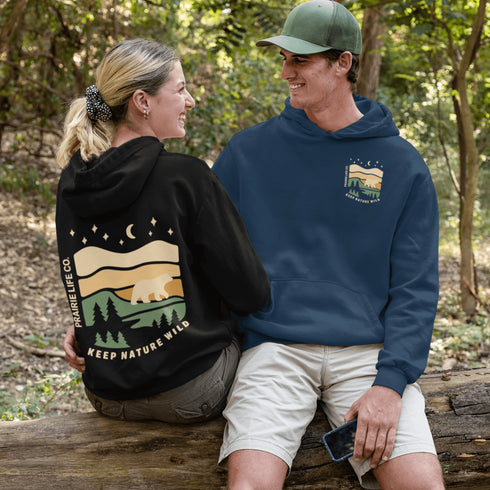 This graphic features a bear silhouette wandering through layered prairie hills, pine forests, and a serene night sky filled with stars and a crescent moon.