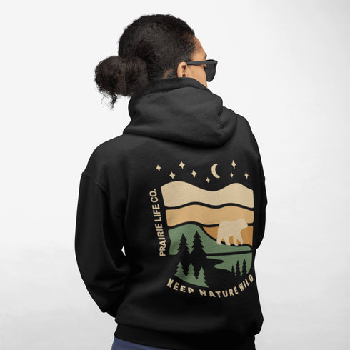 This graphic features a bear silhouette wandering through layered prairie hills, pine forests, and a serene night sky filled with stars and a crescent moon.