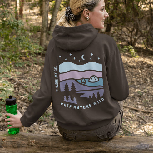 This hoodie features a nature graphic showcasing prairie hills, pine trees, a cozy tent, and a glowing campfire under a crescent moon and star-filled sky.