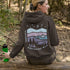 This hoodie features a nature graphic showcasing prairie hills, pine trees, a cozy tent, and a glowing campfire under a crescent moon and star-filled sky.
