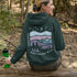 This hoodie features a nature graphic showcasing prairie hills, pine trees, a cozy tent, and a glowing campfire under a crescent moon and star-filled sky.