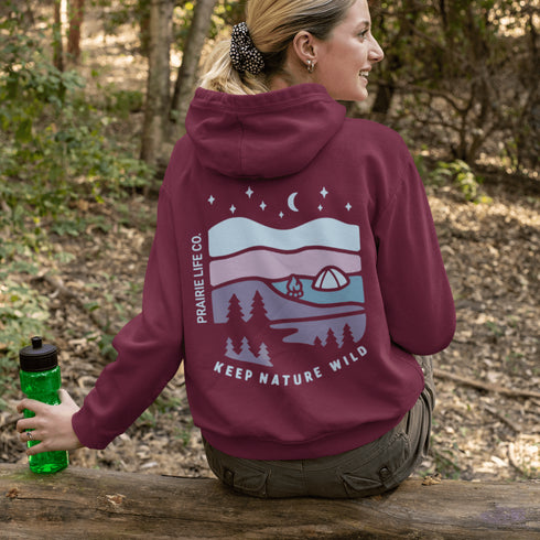 This hoodie features a nature graphic showcasing prairie hills, pine trees, a cozy tent, and a glowing campfire under a crescent moon and star-filled sky.