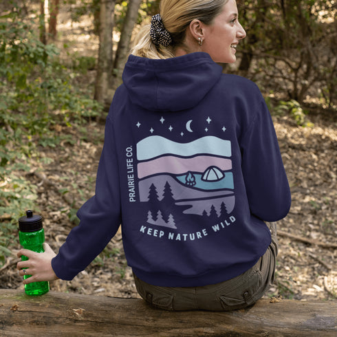 This hoodie features a nature graphic showcasing prairie hills, pine trees, a cozy tent, and a glowing campfire under a crescent moon and star-filled sky.