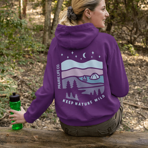 This hoodie features a nature graphic showcasing prairie hills, pine trees, a cozy tent, and a glowing campfire under a crescent moon and star-filled sky.