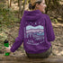 This hoodie features a nature graphic showcasing prairie hills, pine trees, a cozy tent, and a glowing campfire under a crescent moon and star-filled sky.