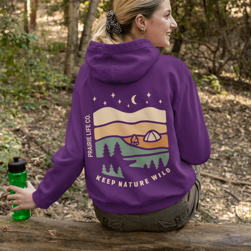 This hoodie features rolling prairie hills, a glowing campfire beside a cozy tent, towering pines, and a star-filled night sky complete with a crescent moon.