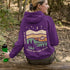 This hoodie features rolling prairie hills, a glowing campfire beside a cozy tent, towering pines, and a star-filled night sky complete with a crescent moon.