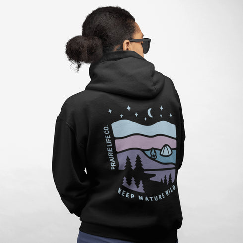 This hoodie features a nature graphic showcasing prairie hills, pine trees, a cozy tent, and a glowing campfire under a crescent moon and star-filled sky.