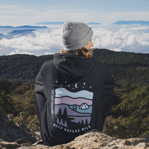 This hoodie features a nature graphic showcasing prairie hills, pine trees, a cozy tent, and a glowing campfire under a crescent moon and star-filled sky.