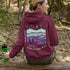 This hoodie features a retro-style landscape graphic with prairie hills, pine trees, a campfire, a tent, and a night sky filled with stars and a crescent moon.