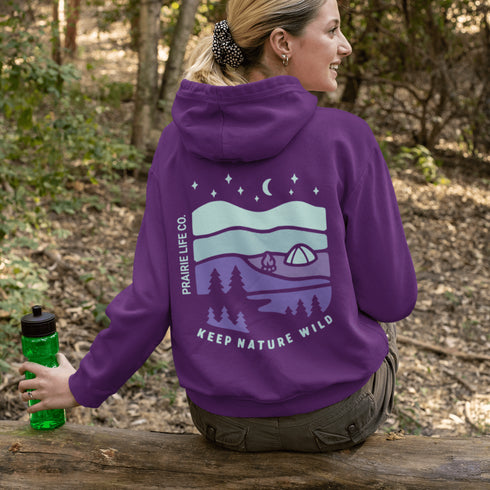 This hoodie features a retro-style landscape graphic with prairie hills, pine trees, a campfire, a tent, and a night sky filled with stars and a crescent moon.