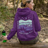 This hoodie features a retro-style landscape graphic with prairie hills, pine trees, a campfire, a tent, and a night sky filled with stars and a crescent moon.