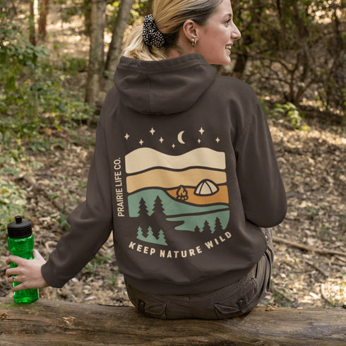 This hoodie features rolling prairie hills, a glowing campfire beside a cozy tent, towering pines, and a star-filled night sky complete with a crescent moon.