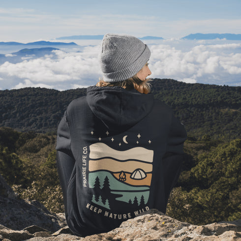 This hoodie features rolling prairie hills, a glowing campfire beside a cozy tent, towering pines, and a star-filled night sky complete with a crescent moon.