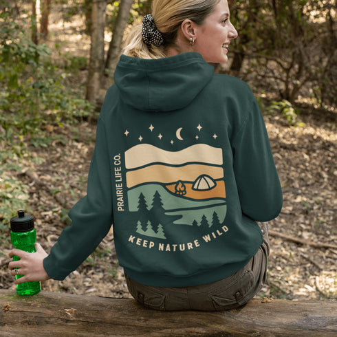 This hoodie features rolling prairie hills, a glowing campfire beside a cozy tent, towering pines, and a star-filled night sky complete with a crescent moon.