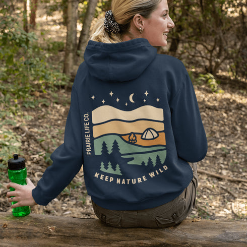 This hoodie features rolling prairie hills, a glowing campfire beside a cozy tent, towering pines, and a star-filled night sky complete with a crescent moon.
