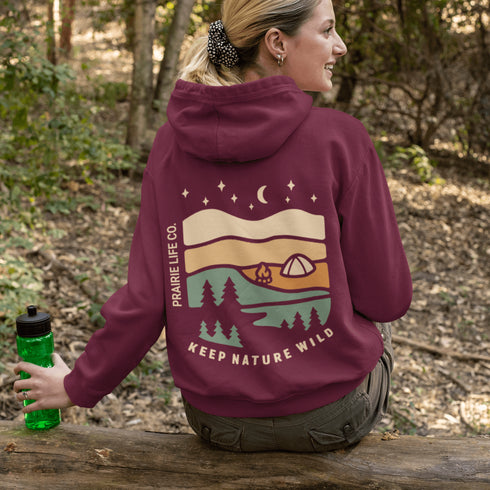 This hoodie features rolling prairie hills, a glowing campfire beside a cozy tent, towering pines, and a star-filled night sky complete with a crescent moon.