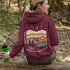 This hoodie features rolling prairie hills, a glowing campfire beside a cozy tent, towering pines, and a star-filled night sky complete with a crescent moon.