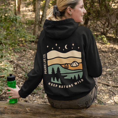 This hoodie features rolling prairie hills, a glowing campfire beside a cozy tent, towering pines, and a star-filled night sky complete with a crescent moon.