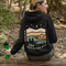 This hoodie features rolling prairie hills, a glowing campfire beside a cozy tent, towering pines, and a star-filled night sky complete with a crescent moon.