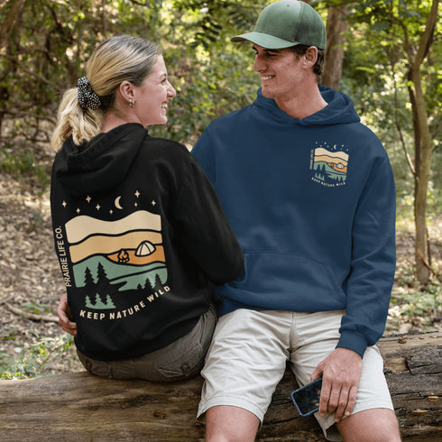 This hoodie features rolling prairie hills, a glowing campfire beside a cozy tent, towering pines, and a star-filled night sky complete with a crescent moon.