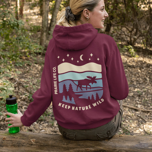 This hoodie features a moose silhouette roaming across prairie hills, pine trees with stars scattered above and a crescent moon glowing softly.